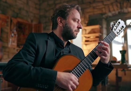 Marcin Dylla podczas występu, zbliżenie na dłonie na gryfie gitary klasycznej, w tle rozmazana scena koncertowa.