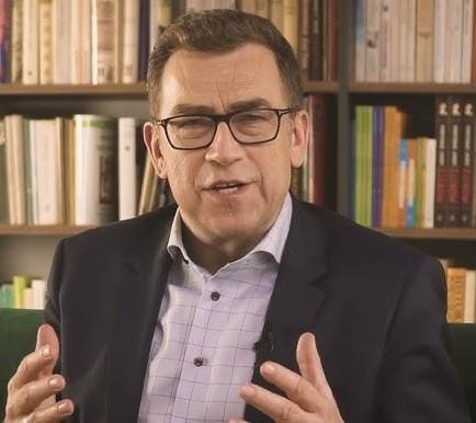 Maciej Orłoś uśmiechnięty, w dynamicznej pozie, w tle nowoczesne wnętrze, odzwierciedlające jego obecne miejsce zamieszkania.