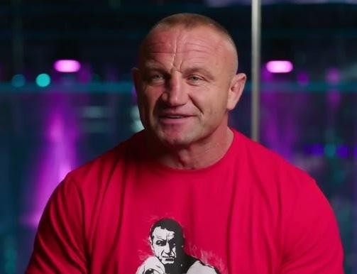 Alt: Mariusz Pudzianowski w klatce MMA, w dynamicznej pozie, prezentujący swoją muskulaturę i determinację przed walką.