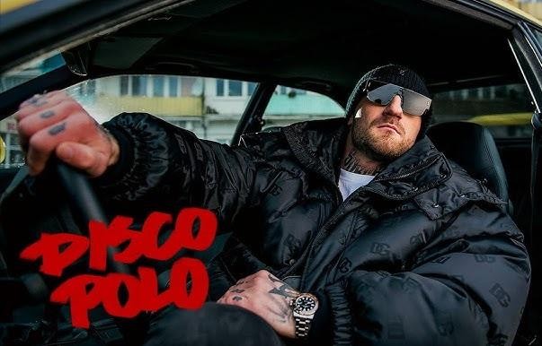 Kizo (Patryk Woziński) podczas występu na scenie lub w studiu nagraniowym, ubrany w streetwear, z elementami muzycznymi w tle.
